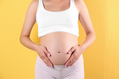 Young pregnant woman holding hands on belly against color background, closeup