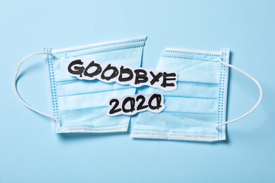 Text Goodbye 2020 and medical face mask on light blue background, flat lay
