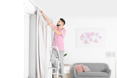 Photo of Young man hanging window curtain in room. Space for text