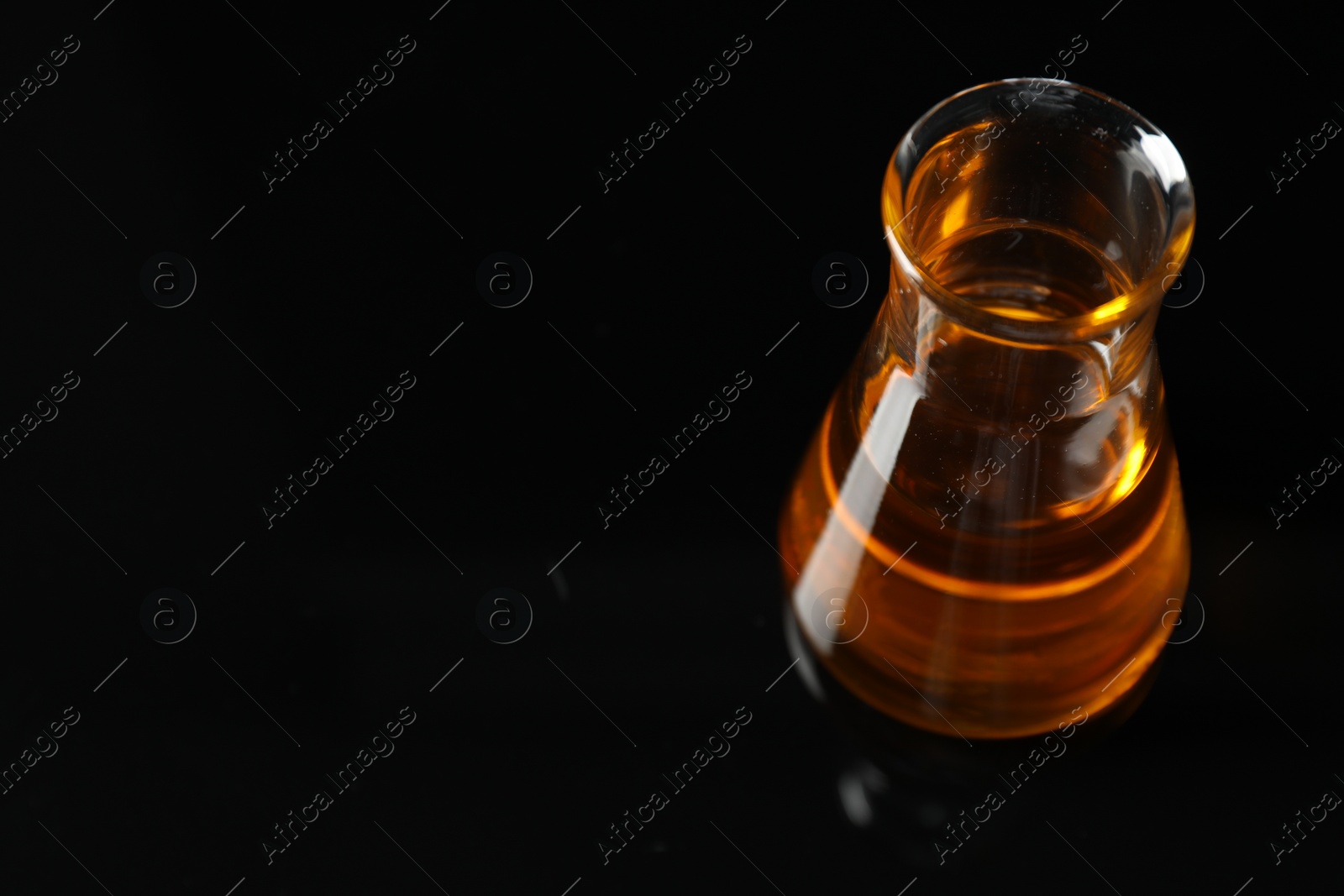 Photo of Glass flask with yellow oil on black background, closeup. Space for text