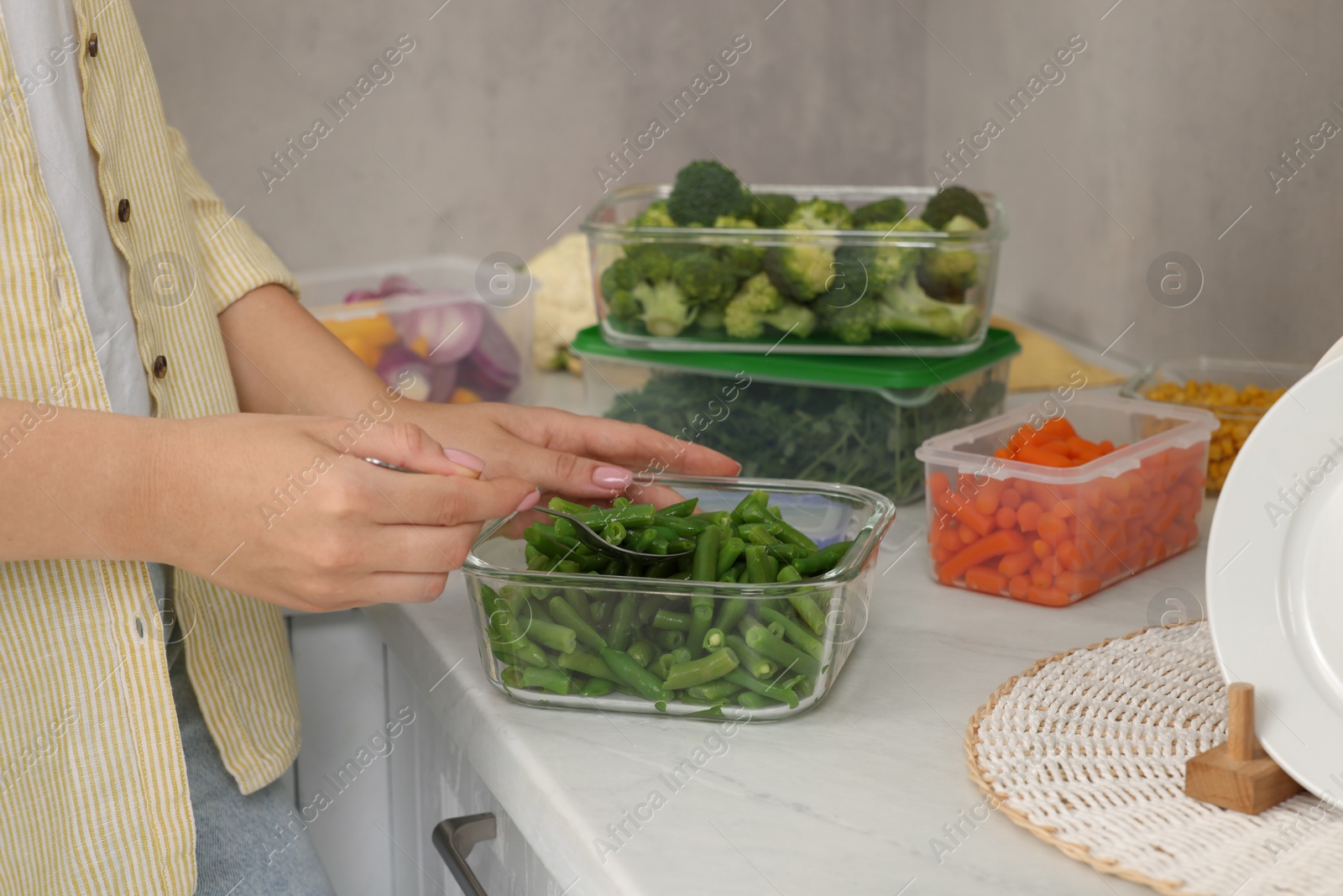 Photo of Woman taking green beans with spoon from glass container at white marble table in kitchen, closeup. Food storage