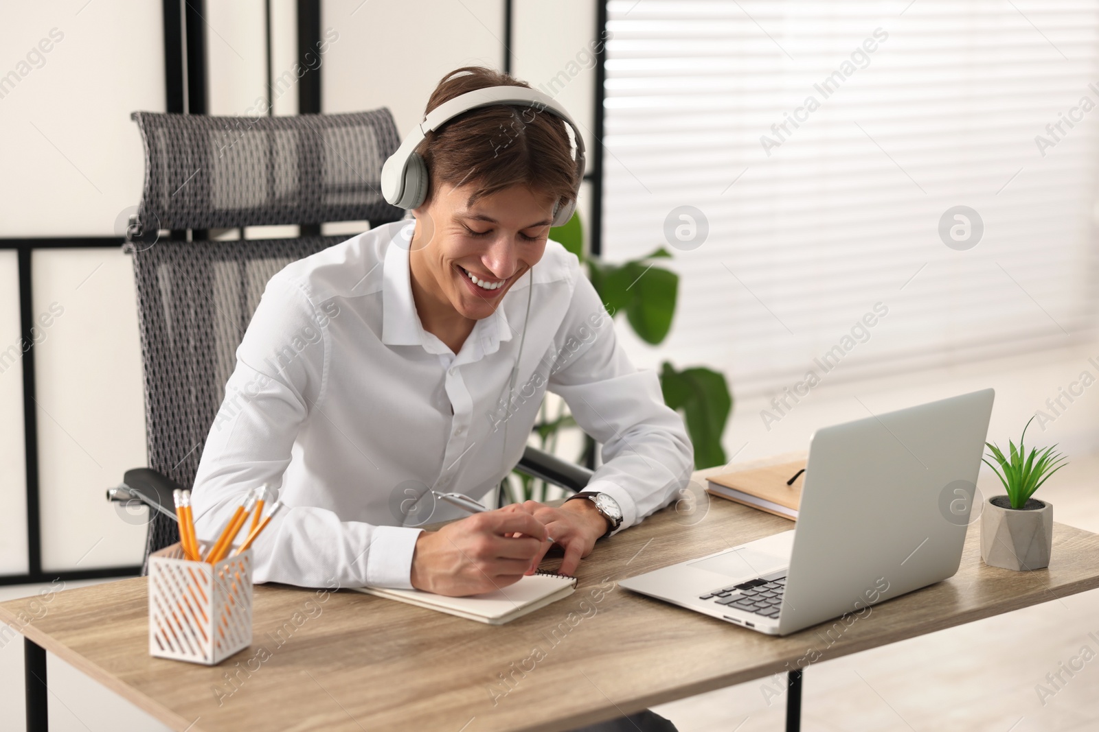 Photo of Man in headphones taking notes during webinar at wooden table indoors