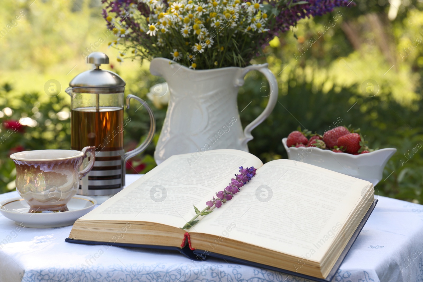 Photo of Open book, tea, ripe strawberries and bouquet of beautiful wildflowers on table in garden