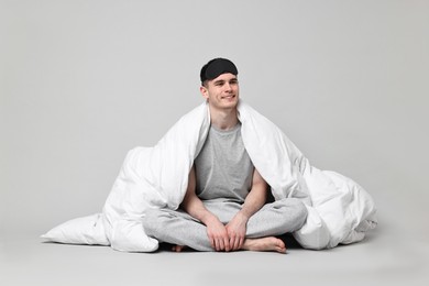 Happy man in pyjama and sleep mask wrapped in blanket on grey background