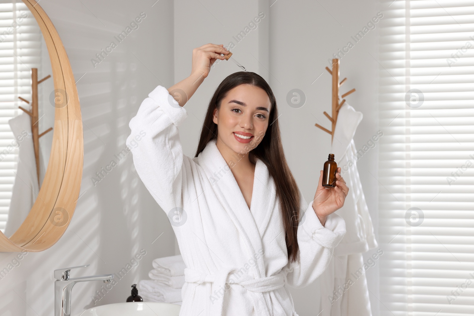 Photo of Happy young woman applying essential oil onto hair roots in bathroom