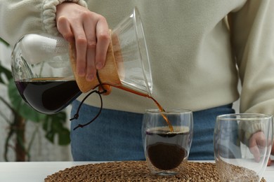 Photo of Woman pouring drip coffee from chemex coffeemaker into glass at table, closeup