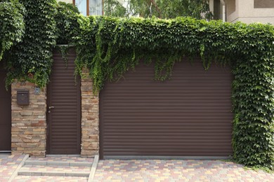 Brown roller shutter door of garage near entrance overgrown with beautiful climbing plants