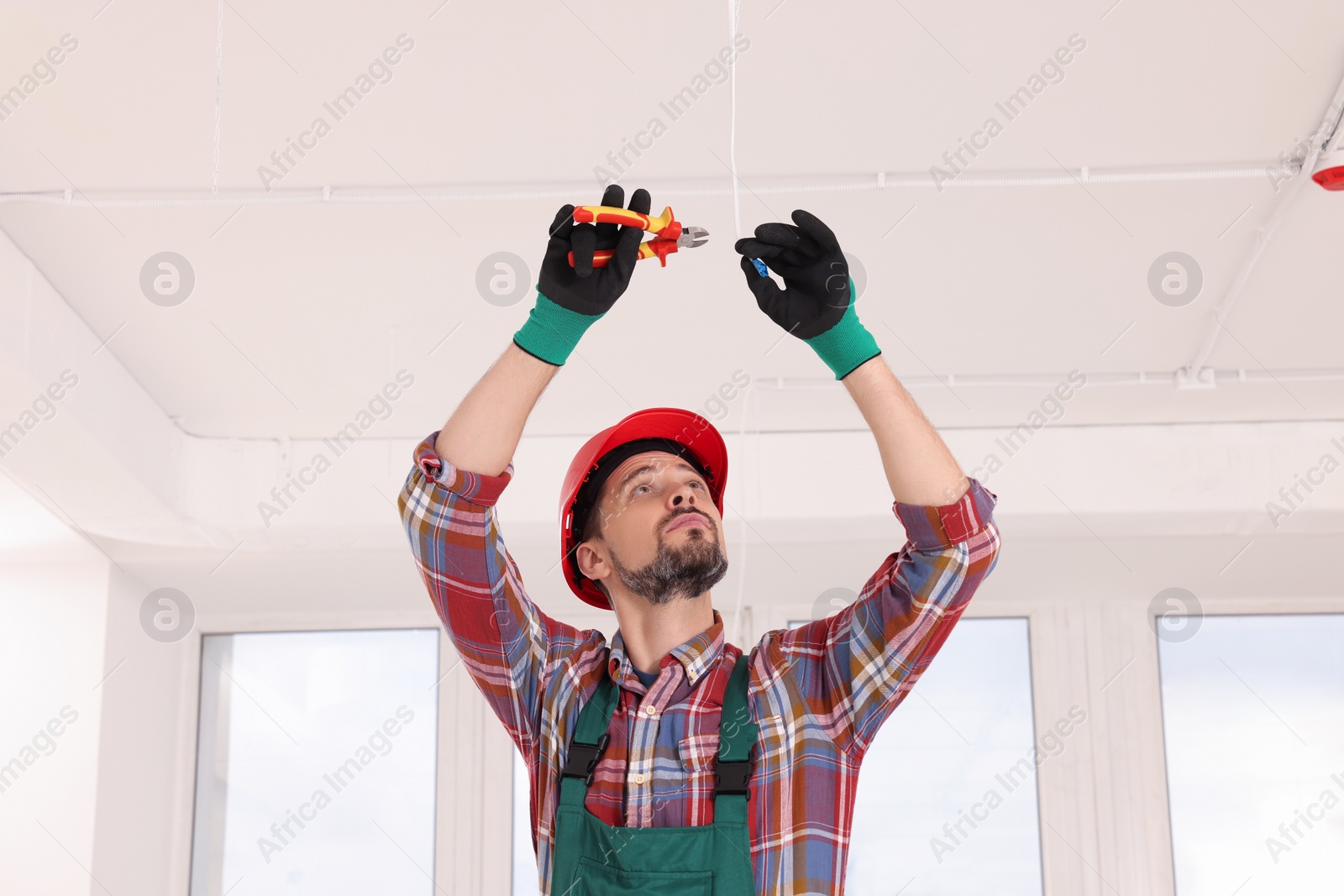 Photo of Electrician in uniform with pliers repairing ceiling wiring indoors