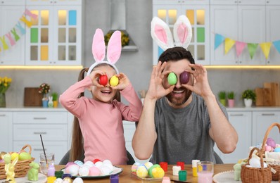 Photo of Father and his cute daughter covering eyes with beautifully painted Easter eggs at table in kitchen