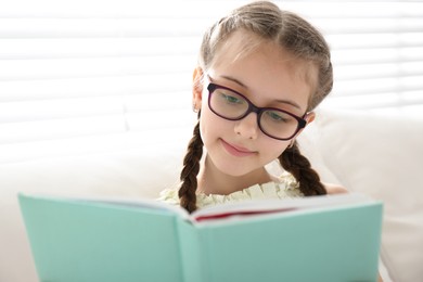 Photo of Cute little girl reading book near window at home