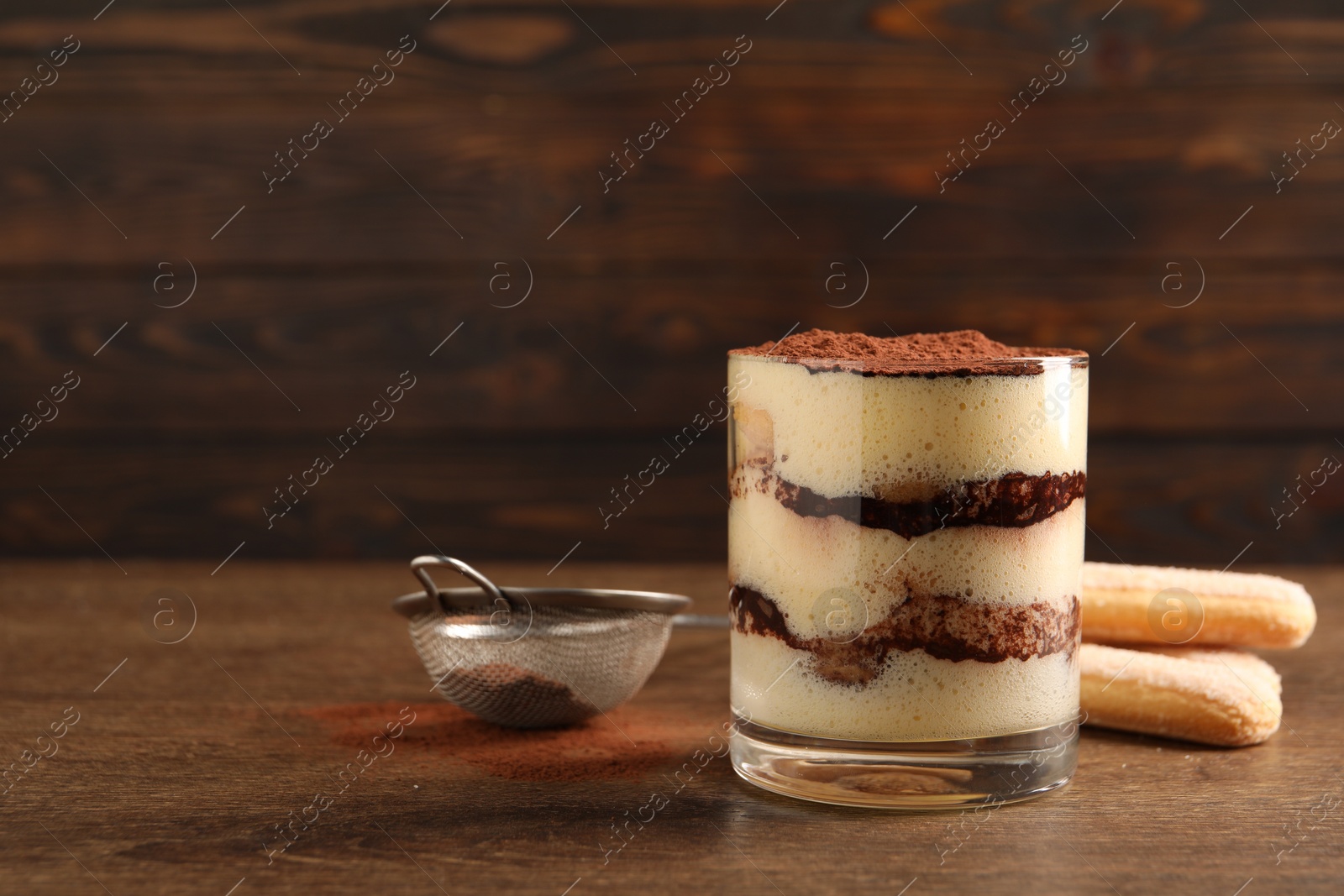 Photo of Delicious tiramisu in glass, biscuits and sieve with cocoa powder on wooden table. Space for text