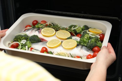 Photo of Woman putting baking dish with raw fish and vegetables into oven, closeup