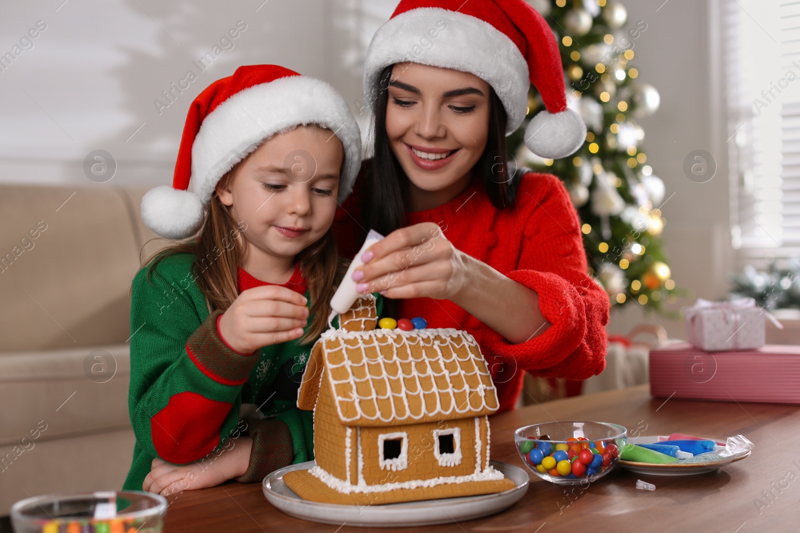 Photo of Mother and daughter decorating gingerbread house at table indoors