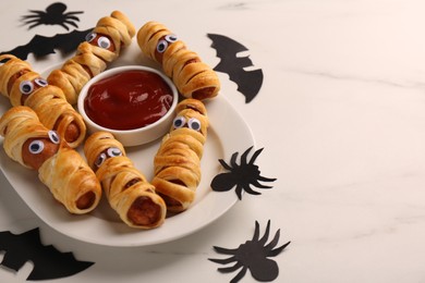 Photo of Plate with tasty sausage mummies, Halloween decorations and ketchup on white table, closeup. Space for text