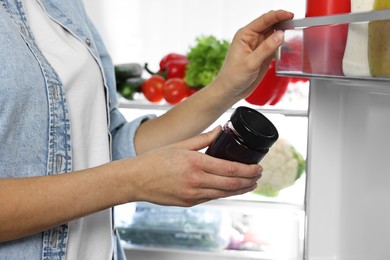 Photo of Young woman taking jar of jam out of refrigerator, closeup