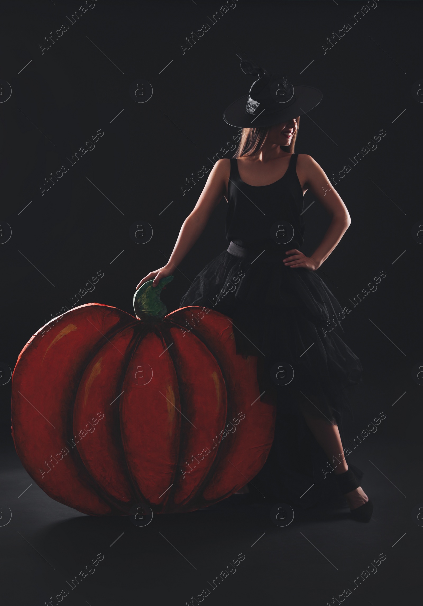 Photo of Young woman wearing witch costume near decorative pumpkin on black background. Halloween party