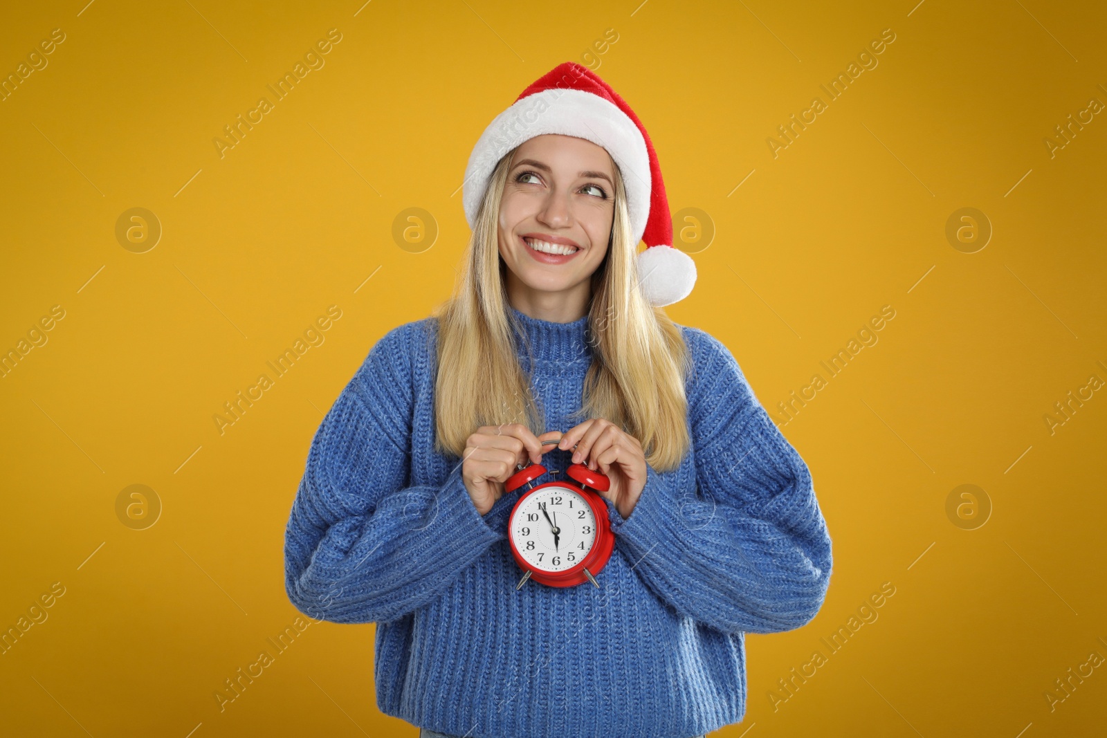 Photo of Woman in Santa hat with alarm clock on yellow background, space for text. New Year countdown