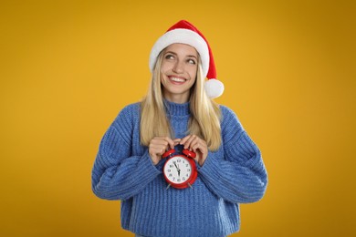 Photo of Woman in Santa hat with alarm clock on yellow background, space for text. New Year countdown