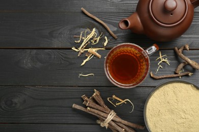 Aromatic licorice tea in cup, teapot, dried sticks of licorice root and powder on black wooden table, flat lay. Space for text