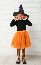 Photo of Cute little girl with candies wearing Halloween costume near white brick wall