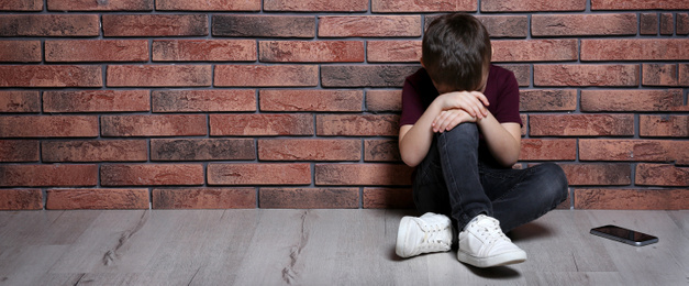 Sad little boy with mobile phone sitting on floor near brick wall, banner design and space for text. Time to visit child psychologist