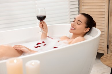 Photo of Happy woman with glass of wine taking bath in tub with foam and rose petals indoors