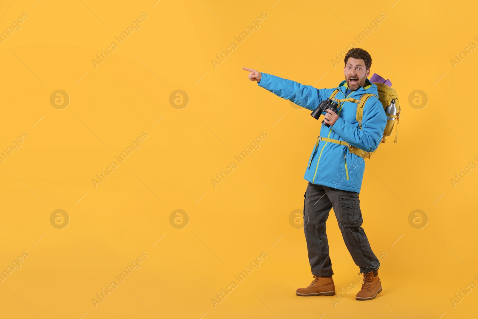 Photo of Emotional man with backpack and binoculars pointing at something on orange background, space for text. Active tourism