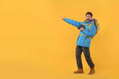 Photo of Emotional man with backpack and binoculars pointing at something on orange background, space for text. Active tourism