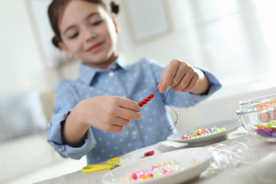 Photo of Little girl making accessory with beads at table indoors, focus on hands. Creative hobby