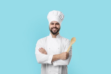 Photo of Happy young chef in uniform holding wooden spoon on light blue background