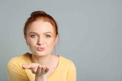 Portrait of young red haired woman blowing kiss on light grey background, space for text