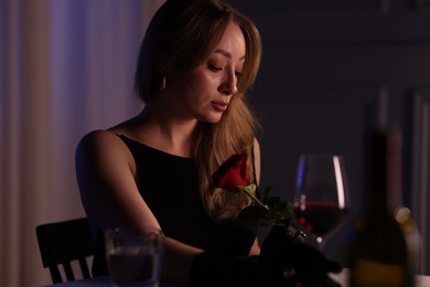 Photo of Elegant young woman with rose and glass of wine at table indoors in evening