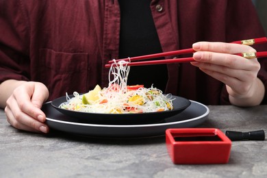 Photo of Stir-fry. Woman with chopsticks eating tasty rice noodles with meat and vegetables at grey textured table, closeup