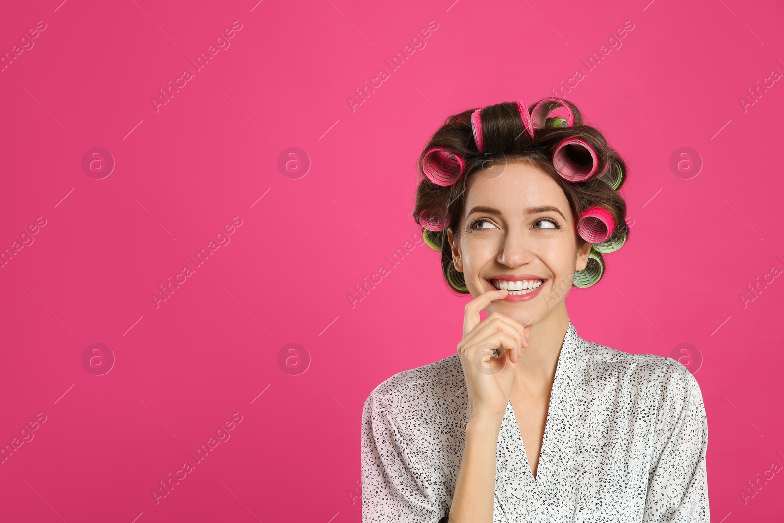 Photo of Beautiful young woman in silk bathrobe with hair curlers on pink background. Space for text