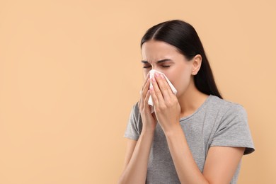 Photo of Suffering from allergy. Young woman blowing her nose in tissue on beige background. Space for text