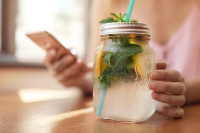 Young woman using mobile phone while drinking tasty natural lemonade at table in cafe. Detox drink