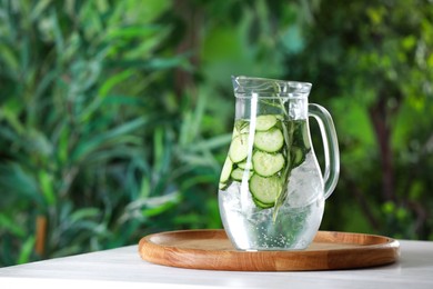 Photo of Refreshing cucumber water with rosemary in jug on white table against blurred green background. Space for text