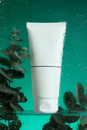 Photo of Tube with moisturizing cream and eucalyptus branches on green background, view through wet glass