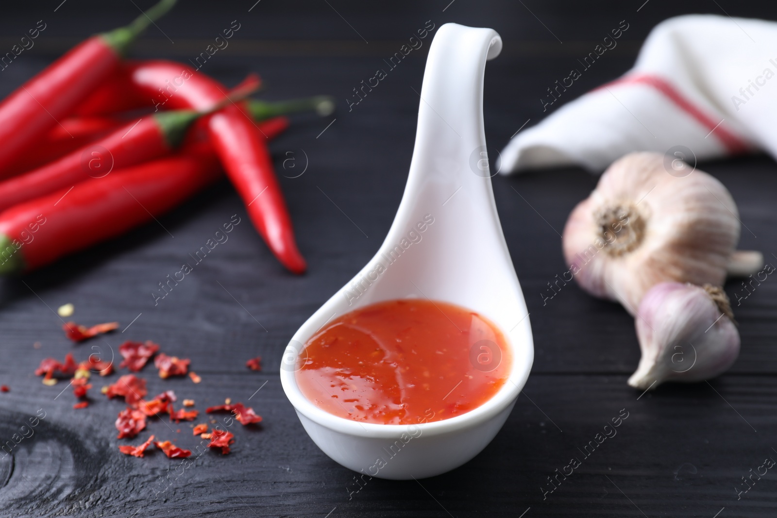 Spicy chili sauce in spoon, peppers and garlic on black wooden table, closeup Photo of Spicy chili sauce in spoon, peppers and garlic on black wooden table, closeup