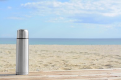 Photo of Metallic thermos with hot drink on wooden surface near sea, space for text