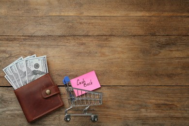 Photo of Card with word Cashback, shopping cart and dollar banknotes in wallet on wooden background, flat lay. Space for text