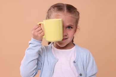 Photo of Happy girl covering eye with yellow ceramic mug on beige background