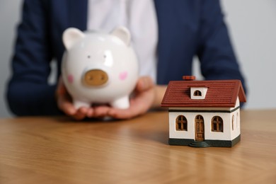 Woman holding piggy bank at wooden table, focus on little house model