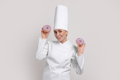 Happy professional confectioner in uniform holding delicious doughnuts on light grey background