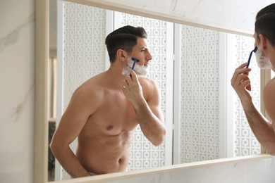 Handsome man shaving near mirror in bathroom