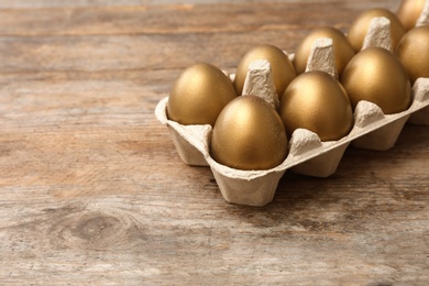 Carton with golden eggs on wooden table, space for text