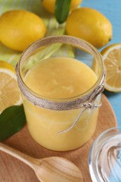 Photo of Delicious lemon curd in glass jar, fresh citrus fruits, spoon and green leaves on table, closeup