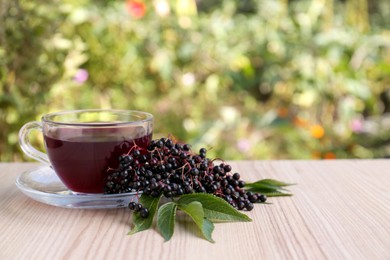 Photo of Glass cup of tasty elderberry tea and Sambucus berries on wooden table outdoors, space for text