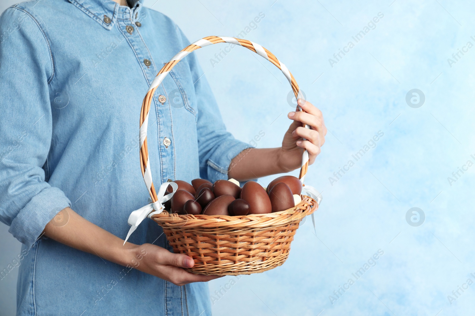 Photo of Woman holding wicker basket with chocolate Easter eggs on color background, closeup. Space for text