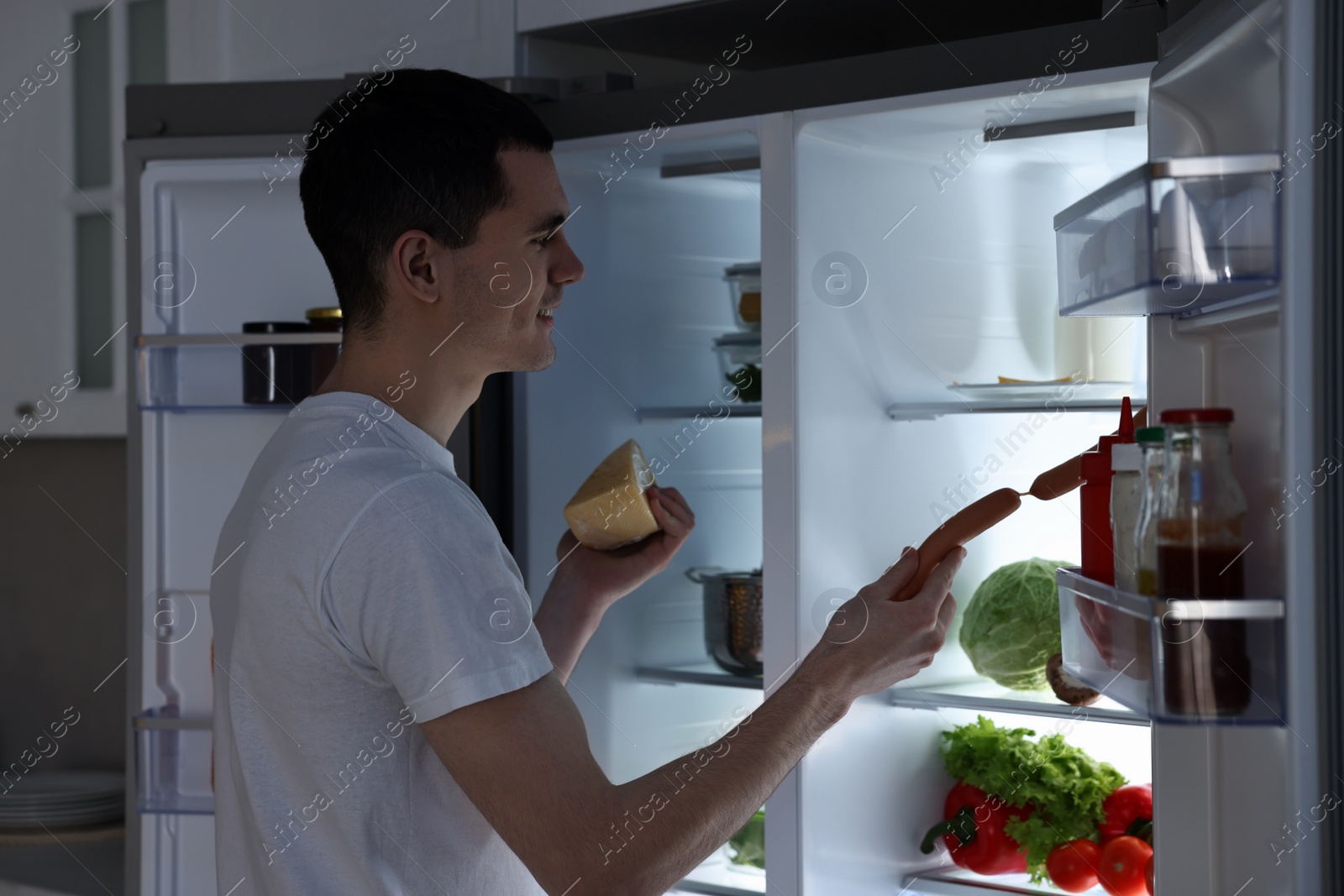 Photo of Man with cheese taking sausages out of refrigerator in kitchen at night